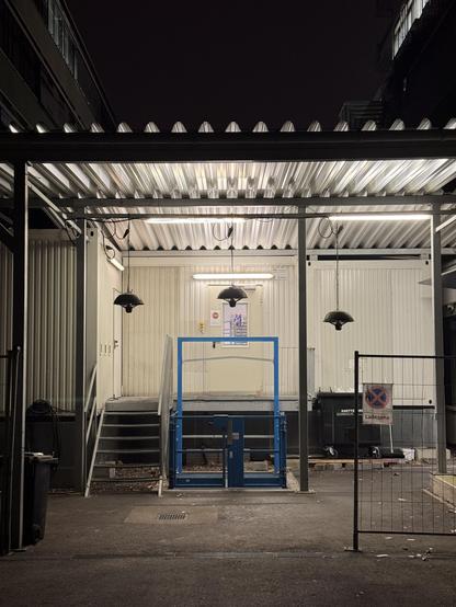 Fabrikhintereingang bei Nacht. Das Tor ist offen, der Ladelift geschlossen, die Neonröhren sind noch an und Hängelampen sind aus.