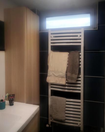 Une fenêtre horizontale étroite en hauteur sur un mur en carrelage noir d'une salle de bain avec un sèche-serviettes blanc sous celle-ci. Sur le côté droit, un lavabo blanc et à sa droite dans le coin un placard en bois clair.