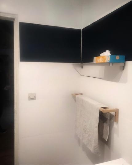 Deux murs d'une salle de bain blancs avec une bande horizontale en carrelage noir avec une étagère fine sur laquelle est posée une boîte de mouchoirs avec un porte-serviettes en bois clair sous celle-ci. Sur le côté droit, un bout de lavabo blanc. À gauche une porte ouverte sur une salle dans le noir.
