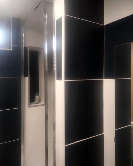 Une douche à l'italienne blanche avec des carreaux de carrelage noir sur les côtés, avec dedans une petite alcôve blanche rectangulaire haute avec le fond en carrelage noir pour poser le savon. Sur la gauche une fenêtre horizontale étroite en hauteur sur un mur en carrelage noir d'une salle de bain.