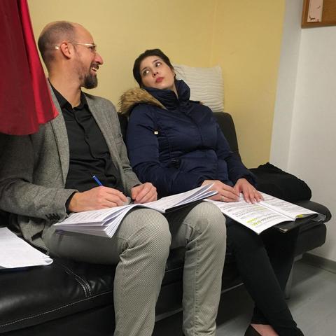 Two people on a black leather sofa in front of a yellow wall look at each other. On the left sits pianist Giacomo Marignani with spectacles, short-cropped hair and beard, wearing a black shirt, grey jacket and grey trousers. On the right, with her black hair pinned up and wearing red lipstick, soprano Megan Marie Hart sits wearing a dark blue puffer coat with faux fur trim. Both have sheets of music on their laps; they are making notes in their scores in Hart's dressing room after a rehearsal for the upcoming world premiere of the song cycle Letters to Fred by composer Bracha Bdil at the Staatstheater Darmstadt on 26 January 2025. Photo: Stefan Romero Grieser, CC-BY-SA 4.0