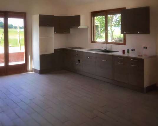 Une cuisine aux murs blancs et au sol en carrelage imitation planches de bois foncé grisâtre au sol, avec sur la droite des meubles de cuisine en bois foncé et une fenêtre avec des meubles hauts en bois foncé de chaque côté et l'évier en dessous. À gauche une double porte-fenêtre qui montre un jardin et un petit muret en brique.