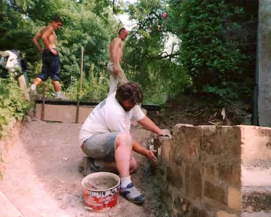 Des travailleurs en arrière plan dans un jardin et au premier plan au milieu Pascal MOREL, dirigeant de J.M.B. Construction, qui monte un muret à droite.