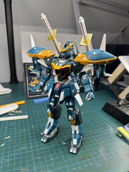 A detailed model of a robotic figure stands prominently on a green cutting mat. The model features blue and orange armor, with large shoulder pads and sword-like accessories attached to its back. Various modeling tools and materials can be seen in the background, indicating a work in progress. 