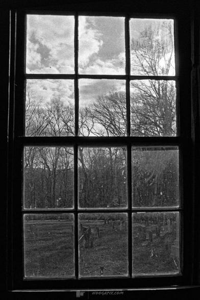 A black-and-white photo taken through a church window, showing a cemetery with gravestones and bare trees under a cloudy sky.