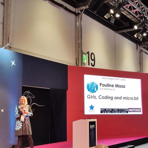 A blonde woman in a floral dress  on stage, giving a talk called Girls, Coding and micro:bit