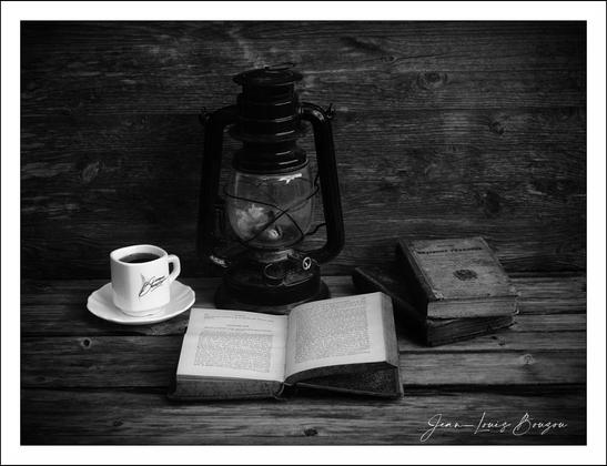 Close-up view of a still life arrangement in monochrome, featuring an antique kerosene lantern, a small cup of coffee, an open book, and a stack of older books on a rustic wooden surface. 
