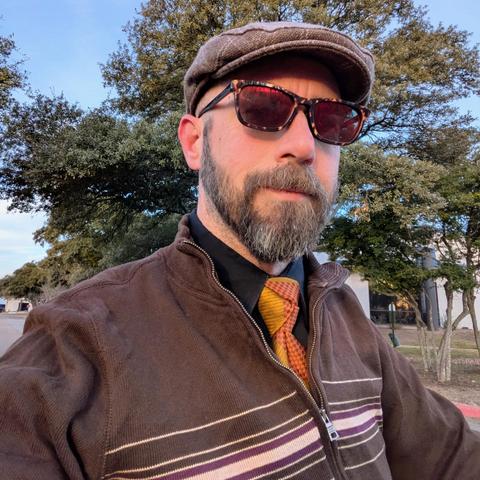 A photo of me, a 46 year old white man with a shaved head, a short cropped brown & grey beard, 

photographed from the chest up in front of some live oak trees in an office park,

wearing

a rectangle glasses with tortoiseshell frames, an olive necktie with orange checkered stripes in a trinity knot on a black dress shirt under a brown quarter-zip sweater with a light brown flat cap
