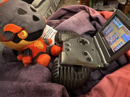 A Charcadet Pokedoll plushie sitting in front of a charcoal coloured Gameboy Advance SP. The screen is displaying the Celadon Game Corner from Pokemon Fire Red. 