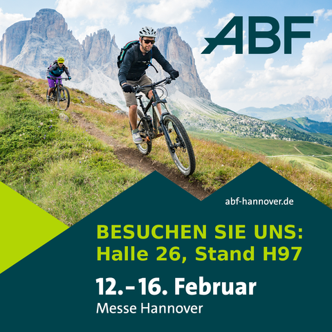 2 Radfahrende fahren einen Wiesenpfad bergab. Im Hintergrund ist hohes Gebirgsmassiv zu sehen mit bewölktem Himmel.