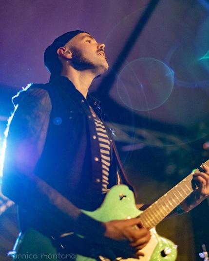 A musician wearing a black cap and a sleeveless jacket plays a light green guitar on stage, with blue and purple lights creating a vibrant atmosphere.