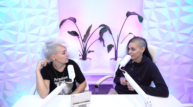 La imagen muestra a dos personas sentadas en un espacio con estética moderna y retrofuturista. Están frente a micrófonos blancos montados en soportes, lo que sugiere que están grabando un podcast.
El fondo es blanco con patrones geométricos en relieve que reflejan luces de colores (principalmente tonos turquesa, fucsia y morado) que crean un ambiente vibrante y llamativo. Detrás de ellas, hay macetas blancas con plantas de hojas grandes, lo que aporta un toque natural al espacio.
Ambas personas parecen relajadas y en conversación. Una de ellas tiene el cabello blanco con estilo despeinado (parecido a una cresta), lleva una camiseta negra con texto y una cadena en el cuello. La otra tiene el cabello rapado en los lados, pelo largo cayendo sobre uno de los lados y viste un suéter azul oscuro. Están mirándose, lo que da una sensación de conexión y naturalidad en su interacción.