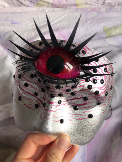 Masque argenté avec des strass noirs et un œil de cyclope rose, réalisé pour une performance drag