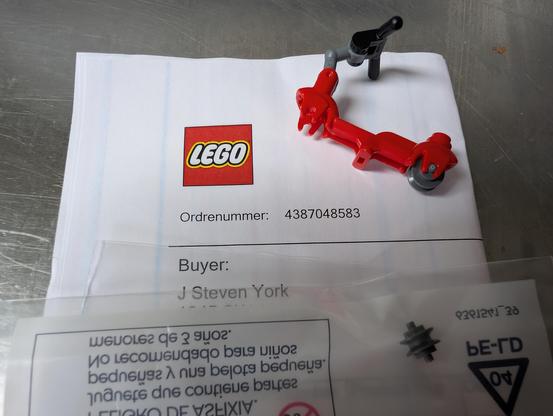 An invoice with a Lego logo on it. In front rests a plastic bag with printing on the reverse, and a tiny gray wheel (it looks more like a pully than a tire) inside. Above it is a tiny Lego scooter, red, with a grey and black handlebar and a missing front wheel.