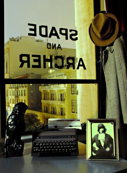 Window with the lettering SPADE & ARCHER a la Sam Spade's office door in THE MALTESE FALCON. Outside we see downtown San Francisco by night. Inside on a table by the window are the falcon, a typewriter and a picture of a woman dressed in 1930s clothing. There is also a coat rack with a coat and hat by the window.