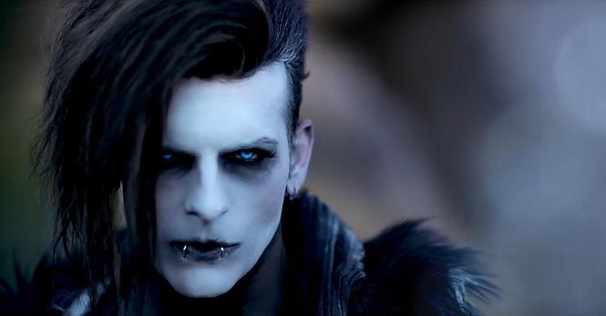 A freeze frame from the music video "Live as Myself" Ben V. has a black dyed side cut with long hair on the left side, he wears white and black makeup and has blazing blue contact lenses. He also has two silver piercings in his lower lip and wears earrings. He is staring in a gloomy way at the viewer.
