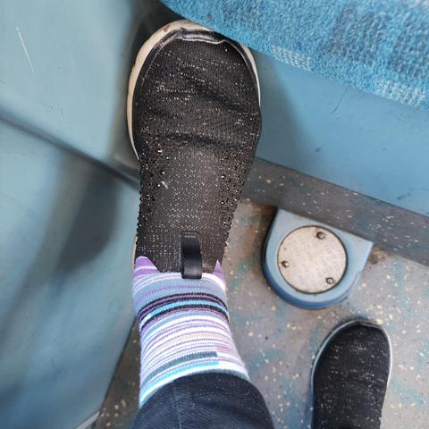 A foot clad in a sparkly black sneaker and a sock with Elizabeth Line striped design rests on the floor of a bus