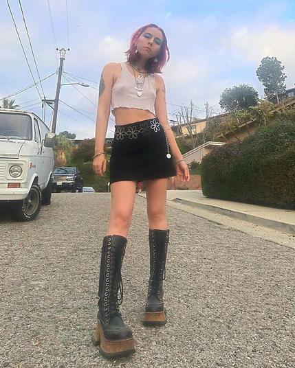The photo features a woman wearing a skirt and boots, set against an outdoor backdrop. The scene includes elements such as the sky and land vehicles, suggesting a casual or fashion-focused moment. Various tags highlight the clothing, footwear, and overall attire of the individual.