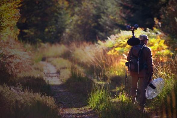 Photo me représentant en forêt, sur un chemin, dans des couleurs automnales et à contrejour, dos tourné, avec du matériel d'enregistrement dans les bras et sur l'épaule.
