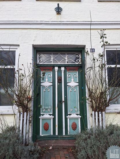 Eine schöne Tür mit vielfältigen Verzierungen in grünen und roten Farben. An den Seiten jeweils winterliche Rosenstöcke und Lavendelbeete.