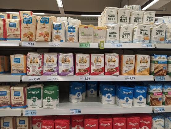 The image shows supermarket shelves stocked with various types of flour in a German grocery store. In the middle shelf, there's a prominent display of Caputo flour, a well-known Italian brand, featuring several varieties in different colored packaging - purple, red, and orange packages labeled "Pizzeria" and "Semola". 