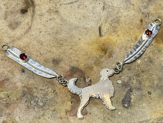 A custom jewelry piece made of silver featuring a dog standing from the side with a smooth surface. On each side of the dog are two long silver feathers that are each adorned with a stone. Work in progress and lightly etched. 