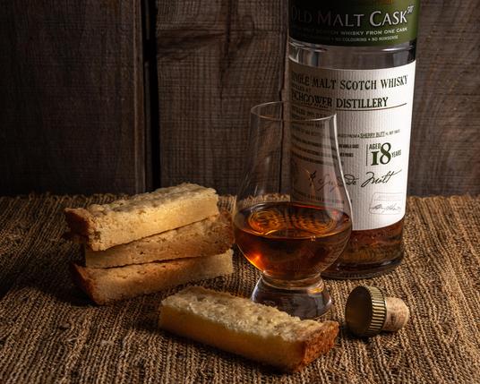 A photograph of a Glencairn glass of whisky, in front of the bottle of Inchgower 18 it was poured from and next to a few pieces of shortbread on a textured burlap surface, with an old wooden wall behind. 
