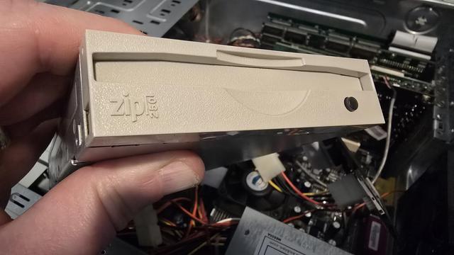 A close-up photograph of a person holding an internal Iomega Zip 250 drive, a vintage storage device from the late 1990s and early 2000s. The beige front panel of the drive features the embossed "Zip 250" logo and a small eject button. In the background, an open computer case with various internal components, including cables, RAM sticks, and a cooling fan, is visible. The lighting suggests a workspace environment for computer maintenance or restoration.