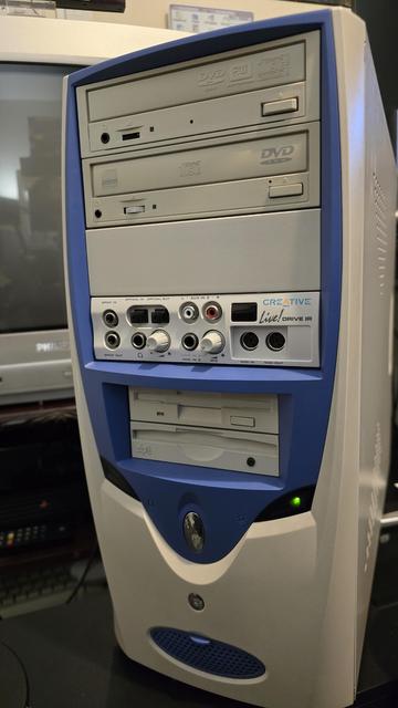 A photograph of a vintage custom-built desktop computer tower with a blue and white case. The front panel features multiple drive bays, including two optical drives (a DVD-RW and a DVD-ROM), a Creative Live! Drive IR audio interface panel with various input and output ports (SPDIF, MIDI, line-in, mic, and auxiliary), and an Iomega Zip drive. Below these components, there is a circular power button with a reflective surface and a small green LED indicator. In the background, an old Philips CRT monitor is visible, along with a partially seen PlayStation console and a keyboard, suggesting a retro computing or gaming setup.