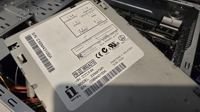 A close-up photograph of an Iomega Zip 250 ATAPI internal drive, showing the label with technical details. The label includes the model number (Z250ATAPI), serial number (S/N: C5BM43100A), power requirements (+5V, 0.8A), and various certification marks (FCC, CE, UL). It also contains jumper settings for Master, Slave, and Cable Select, as well as the manufacturing details, indicating it was assembled in Malaysia. In the background, an open computer case with internal components and cabling is visible, suggesting a repair or restoration process.