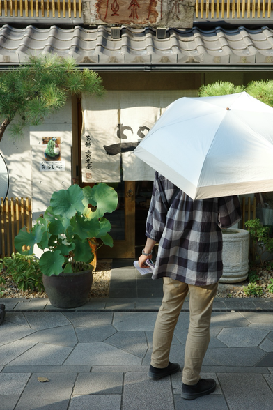 A person with an umbrealla to protect from the strong sun standing o  front of a storefront of a japanese café in Uji, Japan. It is summer and there are multiple potted plants standing in the sun i  front of the café.