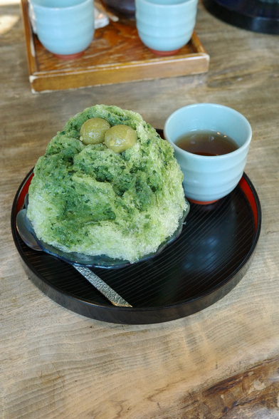 A portion of shaved ice and green tea served on a wooden platter. It is covered with japanese matcha sauce and two mochi are placed on top.