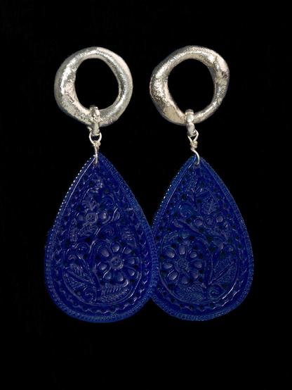 A pair of intricately designed earrings featuring a vibrant blue teardrop shape with floral patterns, suspended from silver circular hooks. The background is black, enhancing the earrings' colors.