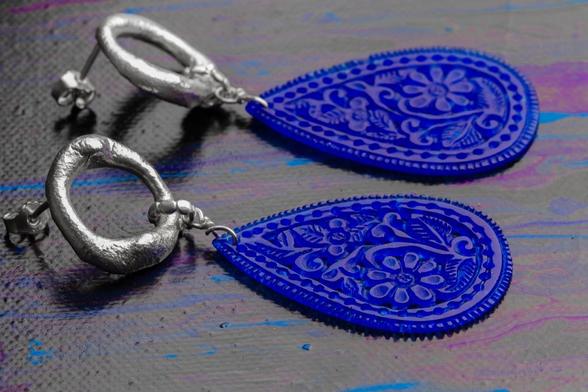 A pair of artistic earrings featuring silver hoops and deep blue teardrop-shaped pendants with intricate floral designs. The background has a textured surface with vibrant colors.