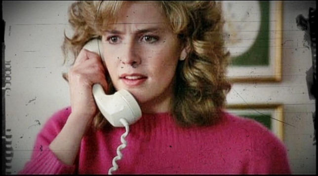 In a still image from the movie "Adventures in Baby Sitting," Elizabeth Shue has a white phone receiver to her ear. She is standing in an interior setting with framed pictures on the wall behind her.  She is wearing a soft pink sweater and her softly curled brown hair falls to her shoulders.