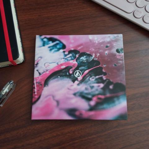 Último álbum de Linkin Park, "From Zero", en formato CD. Estuche estilo digifile cerrada sobre un escritorio, mostrando la carátula. El arte de la carátula es abstracto, realizado sin CGI, mezclando pintura líquida y tomando fotografías. Está en colores rosado, blanco y negro. Al medio se aprecia el logo de Linkin Park en color blanco, con las iniciales "LP" encerradas en un círculo.