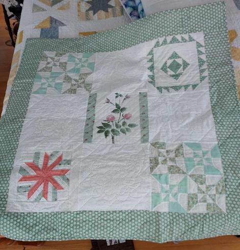 a quilt, it has a thick green and white checkered floral border, in the center is a white square plane, in each corner is a different geometric design done in blues, light greens, and one with light red, a wheel-type design in the lower left corner 
