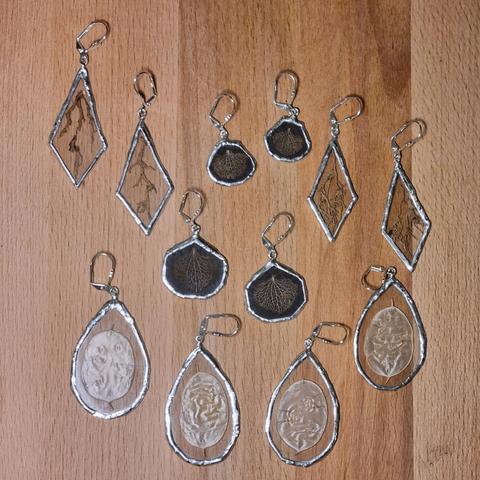 Some handmade stained glass pairs of earrings. Between the glass planes are lunaria discs, plants roots, and skeleton hydrangea petals. The latter have a black background so they stand out more on the skin.