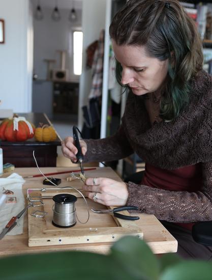 A craftswoman works at her desk, she's soldering stained glass pendants with an iron. Her hair is half dark forest green, half dark blonde.