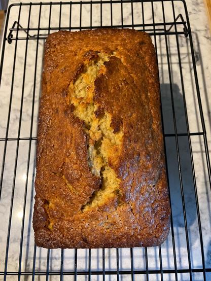 Simple loaf of banana bread atop a baking rack. The loaf has hints of orange and yellow within a dark golden brown top. 