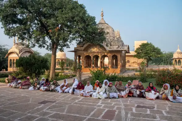 A line of women dressed in white sit on the ground with temple buildings in the background. 