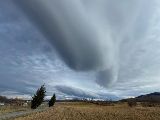 This image shows a landscape with an expansive sky featuring large, elongated lenticular clouds. These clouds have an unusual, smooth, lens-like shape. The scene includes a rural area with fields, a few trees, and distant hills. The lighting appears overcast, contributing to a dramatic and serene atmosphere.
