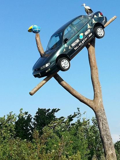 Bildbeschreibung von KI:
Das Bild zeigt eine ungewöhnliche Installation, bei der ein dunkelgrünes Auto schräg auf einem großen, baumartigen Holzgestell platziert ist. Das Auto trägt Aufschriften mit der Werbung für den "Vogelpark Marlow" sowie eine Telefonnummer. Auf dem Dach des Autos sitzt ein dekorativer Storch, und vorne an einem der Holzstämme ist ein Modell eines Vogelschnabels angebracht. Im Hintergrund ist ein klarer blauer Himmel sowie grüne Büsche und Bäume zu sehen.
