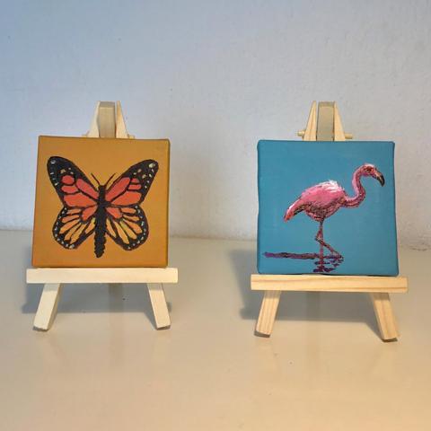 Two small paintings, each on a little easel. Left an orange butterfly, right a pink flamingo standing in a lake.
