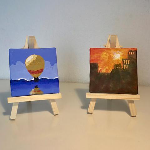 Two small paintings, each on a little easel. Left a hot air balloon over water, right a cityscape with a setting sun.