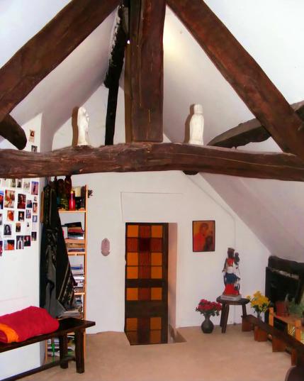 Une chapelle aux murs blancs et à la moquette beige, sous des combles, avec poutres apparentes en bois foncé, une porte avec des vitraux. Il y a plusieurs statues et icônes religieuses et des bouquets de fleurs.  Sur la gauche un banc avec des couvertures et des photos au murs.