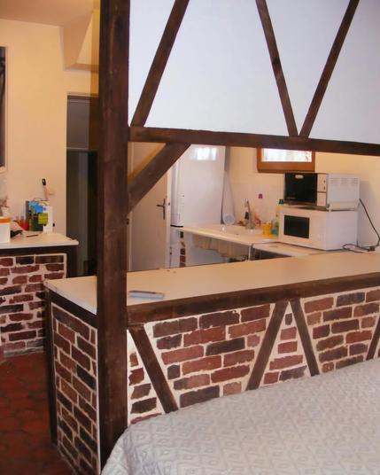 Une cuisine américaine ouverte avec un plan de travail au sous bassement en brique avec effet colombages en bois foncé, surplombé d'un demi-mur blanc avec colombages en bois foncé. Le sol est en tomettes rouges. On voit l'évier sous une fenêtre, un micro-ondes et un mini-four à sa droite. À gauche un second plan de travail identique. Au fond une porte ouverte sur une pièce sombre.