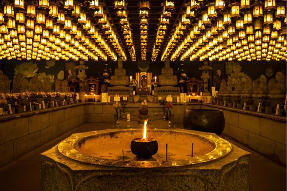 A dimly lit temple interior featuring hundreds of glowing golden lanterns suspended from the ceiling, creating a warm and serene ambiance. In the foreground, a stone basin with a small flame at its center adds a focal point of light. The background showcases statues of Buddhist deities and ornate decorations, emphasizing the sacred and peaceful atmosphere of the space.