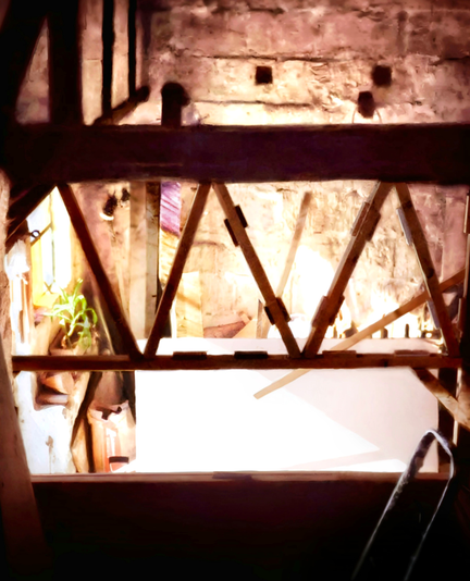 Une pièce en vieille pierre, traversée de poutres en bois foncé avec un demi-mur en dessous. Sur le côté gauche le mur avec des colombages et une fenêtre de vitraux qui illumine la pièce avec des couleurs jaunes et violettes. Il y a des sacs de ciment au sol sous la fenêtre ainsi qu'une plante en pot. Au premier plan à droite il y a un escabeau et au fond des planches diverses.