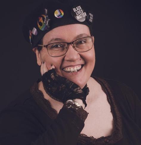 Foto: Amalia trägt eine Brille und eine schwarze Baskenmütze mit mehreren Buttons, darunter "Black Lives Matter" und die Inter* Progressive Pride Farben. Sie/they lächelt den Betrachter an, trägt einen schwarzen Rüschenhandschuh und hat schwarz lackierte Fingernägel, außerdem ein schwarzes Shirt mit Rüschen am Saum. Der Hintergrund des Fotos ist schwarz.